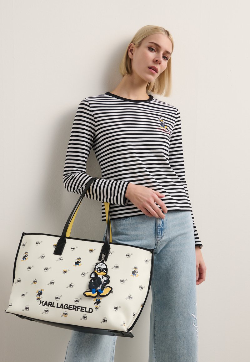 Witte tote bag met zwarte accenten, voorzien van cartoonontwerpen, een gele schouderband en de tekst "KARL LAGERFELD". Model draagt een gestreept shirt.