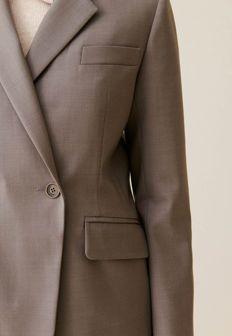 Brązowa marynarka z pojedynczym zapięciem na guzik, wyraźnie wyrzeźbionymi ramionami i kieszenią z przodu. Gładki materiał o subtelnej fakturze i dopasowany krój.