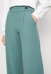 Miętowe spodnie o wysokim stanie i szerokich nogawkach z czarnymi guzikami w talii, noszone z białą bluzką z długim rękawem, ręka w kieszeni.