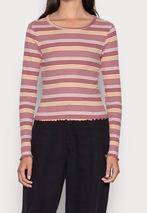 Woman wearing a long-sleeve, fitted top with horizontal pink, beige, and white stripes and black pants against a plain background.
