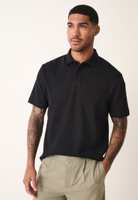 Polo noir à manches courtes avec un motif texturé. Il présente un col classique et une patte à trois boutons. Porté avec un pantalon beige clair.