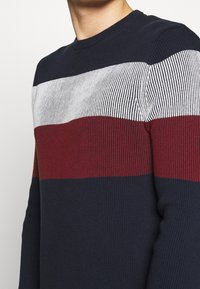 Mörkblått stickat tröja med horisontella ränder i röd, vit och mörkröd. Har rundad halsringning och en strukturerad ribbad mönster.