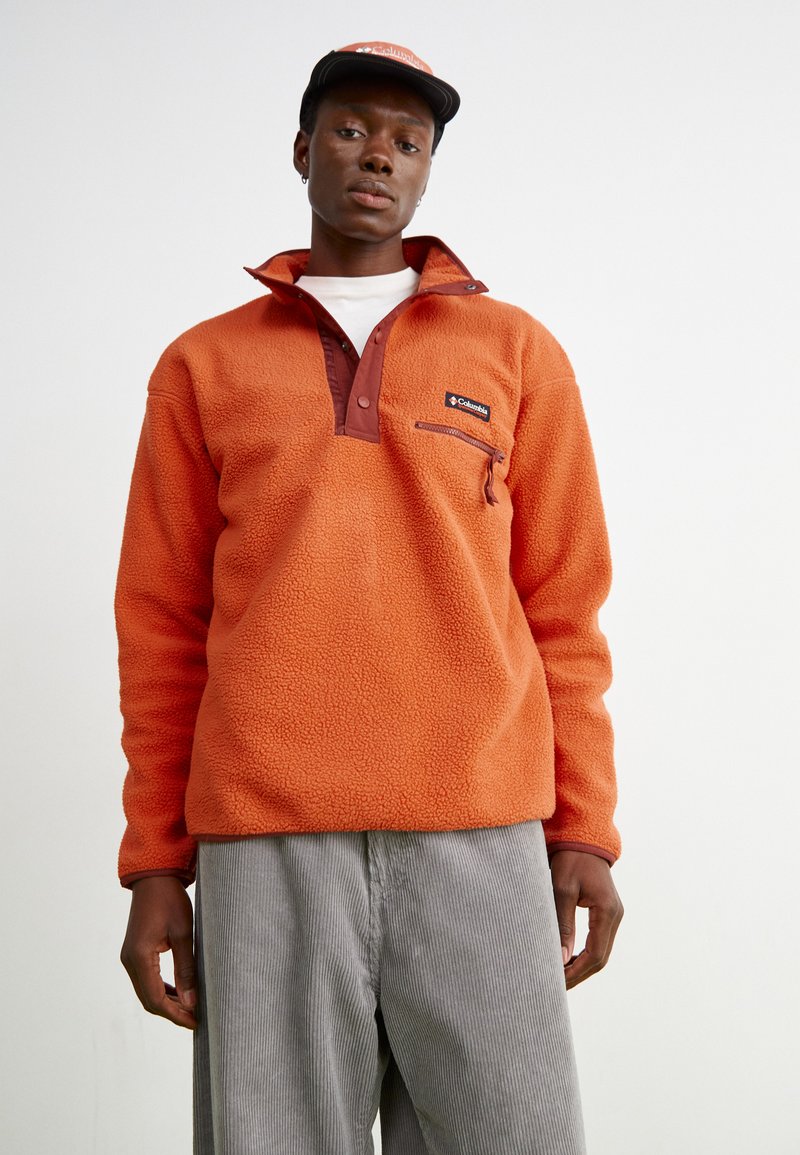 Pullover in pile arancione con colletto alto e zip, caratterizzato da una patta bordeaux a contrasto e una tasca frontale, con texture e vestibilità morbida.