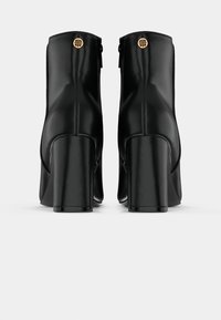 Pár černých kožených kotníkových bot s blokovými podpatky a zlatými kulatými logovými akcenty na zadní části, zobrazených ze zadního pohledu na bílém pozadí.