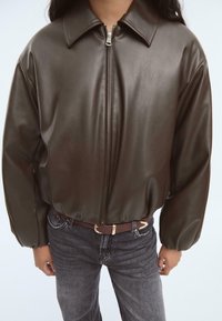 Braune Lederjacke mit Reißverschluss, weitem Kragen, lockerer Passform und voluminösen Ärmeln, kombiniert mit grauen Jeans und einem braunen Gürtel.