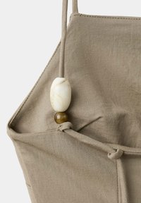 Sac fourre-tout en tissu beige avec une lanière à cordon nouée mettant en valeur une grande perle ovale crème et une petite perle ronde marron.