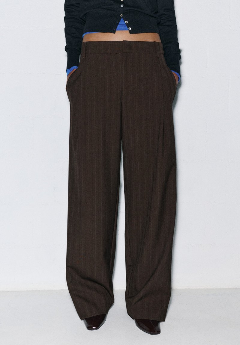 Personne portant un pantalon large marron foncé taille haute à fines rayures, mains dans les poches, associé à un top court noir et des chaussures foncées.