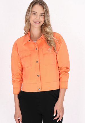 Junge Frau mit langen, welligen blonden Haaren lächelnd, trägt eine leuchtend orangefarbene Jacke mit Knöpfen und schwarze Hosen vor einem einfarbigen Hintergrund.