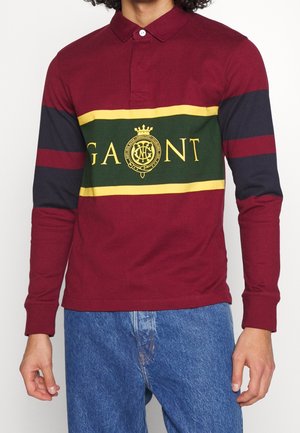 Homme portant un polo manches longues bordeaux avec des rayures horizontales vertes et jaunes et un logo jaune « GANT » sur la poitrine, associé à un jean bleu.