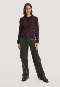 Burgundin värinen neulecardigan, jossa on pyöreä kaula-aukko ja kuusi nappia, yhdistettynä korkeavyötäisiin vihreisiin corduroy-housuihin ja tummiin kenkiin. Sujuva rakenne.