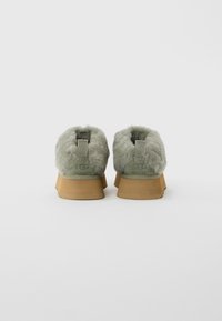 Pair of green suede fur-lined UGG boots viewed from the back, with brown rubber soles on a plain white background.