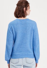 Pull en tricot bleu à manches longues, avec un col rond et un ourlet côtelé. Tissu doux et texturé avec une coupe décontractée, associé à un jean clair.