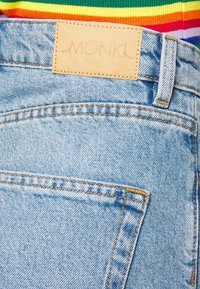 Jeans de mezclilla azul claro con cintura alta, que presentan un parche de cuero beige etiquetado "MONKI" y detalles de costura amarilla en contraste.