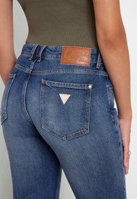 Kõrge vöökohaga sinised stretch teksad, millel on tekstuuriga viimistlus, tagaküljel tan-kollane nahkplekk ja taskul kolmnurkne logo.