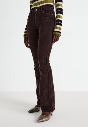 Personne portant un pantalon en velours côtelé brun foncé, un pull rayé à manches longues vert, noir et blanc, et des talons métalliques pointus.