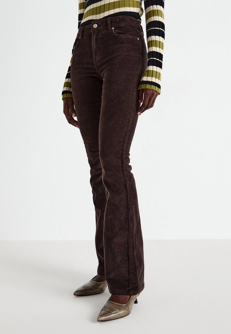 Personne portant un pantalon en velours côtelé brun foncé, un pull rayé à manches longues vert, noir et blanc, et des talons métalliques pointus.