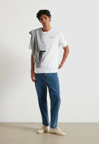 Witte katoen t-shirt met de tekst "FARAH", gecombineerd met blauwe spijkerbroek en beige sneakers. Grijze sweatshirt over de linkerarm gedrapeerd.