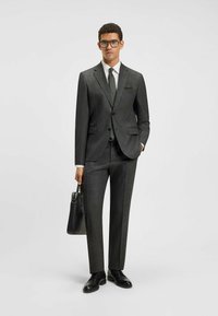 Completo grigio scuro con un motivo a quadri; giacca monopetto, vestibilità aderente, camicia bianca e cravatta a pois. Scarpe nere e borsa.