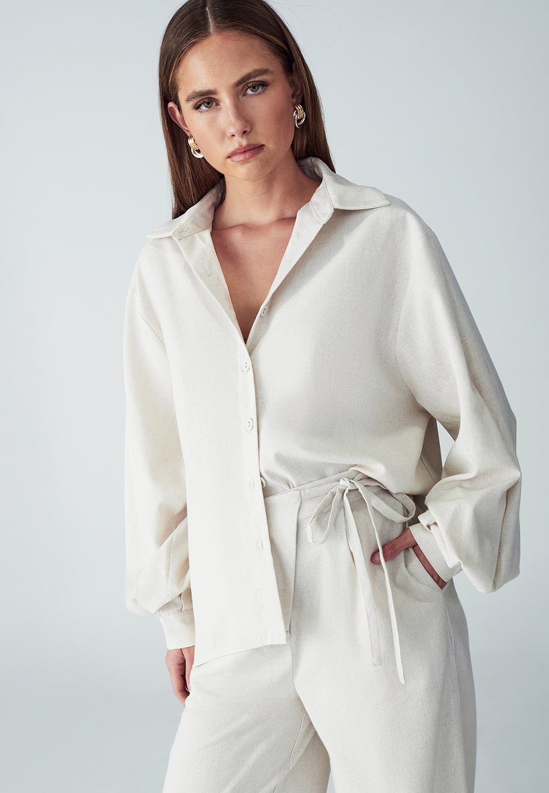 Camicia lunga a maniche con colletto di colore crema, con bottoni frontali e vestibilità morbida, abbinata a pantaloni a vita alta coordinati con dettaglio da annodare.