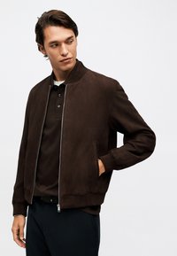 Νέος άνδρας που φοράει σκούρο καφέ σουέτ μπουφάν τύπου μπομπέρ πάνω από μαύρο πόλο πουκάμισο, με το ένα χέρι στην τσέπη, κοιτάζοντας προς τα αριστερά του.