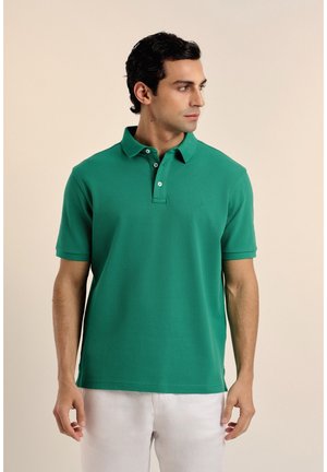 Hombre vestido con camisa tipo polo de manga corta verde y pantalones blancos, mirando hacia su derecha frente a un fondo beige liso.
