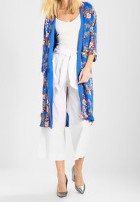 Kimono azul con estampado floral y mangas anchas, superpuesto sobre una blusa blanca y culottes blancos. Los zapatos de tacón plateados completan el conjunto. Textura suave.