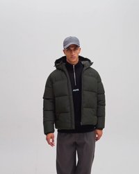 Junger Mann, der eine dunkelgrüne gesteppte Jacke mit Kapuze, ein schwarzes Zip-up-Oberteil, eine graue Kappe und lockere graue Hosen trägt, vor einem schlichten Hintergrund stehend.