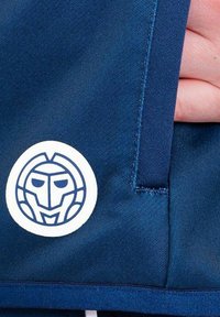 Tessuto navy con una finitura testurizzata, caratterizzato da un logo circolare bianco con un design di faccia stilizzato sul lato di una tasca del capo.