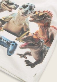 Vit skjorta med färgglada dinosaurtegningar som bär solglasögon, tillsammans med en blå spelkontroll. Rolig design med humoristiska inslag.