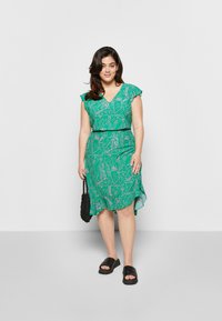 Vestido verde com decote em V e mangas com babados, apresentando um padrão floral em rosa claro. A bainha é assimétrica. Usado com sandálias pretas e uma mala.