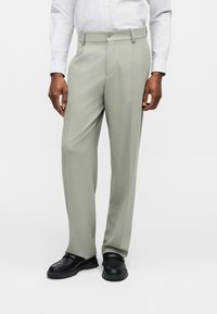 Άνδρας με ανοιχτό γκρι, ραμμένο παντελόνι, λευκό πουκάμισο και μαύρα δερμάτινα μοκασίνια, στέκεται μπροστά σε λευκό απλό φόντο.