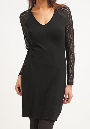 Femme portant une robe noire longueur genoux avec un décolleté en V et des manches longues en dentelle, debout devant un fond blanc uni.