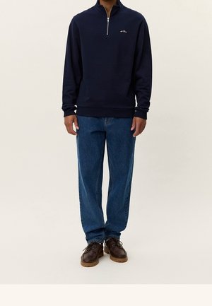 Mand iført mørk marineblå sweatshirt med kvartlynlås, blå straight-leg jeans og brune lædersko står mod en ensfarvet baggrund.