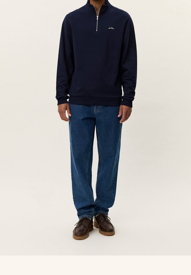 Homme portant un sweat-shirt bleu marine foncé à fermeture éclair quart, jean droit bleu et chaussures en cuir marron, debout sur un fond uni.