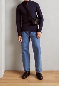 Uomo con maglione blu navy con cerniera, jeans azzurri, stivali neri e borsa a tracolla nera in piedi contro un muro grigio su pavimento in legno.