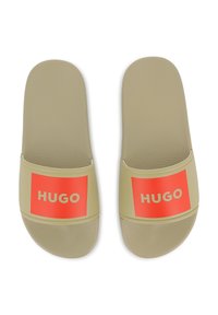 Μπεζ λαστιχένια σανδάλια σαγιονάρας με φαρδιά ταινία που φέρει ένα κόκκινο τετράγωνο και λευκό κείμενο "HUGO." Υφασμένο πέλμα για άνεση.