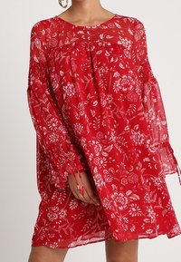 Robe florale rouge avec des manches longues en voile, présentant une coupe ample et un froncé au niveau du col. Motifs floraux dans des tons blancs et roses.