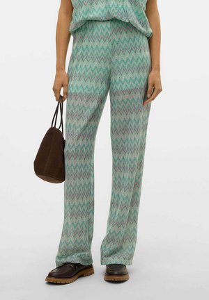 Pantaloni a zigzag verde menta realizzati in tessuto leggero, abbinati a una maglietta ampia e a una borsa marrone, indossati con scarpe marroni scure.