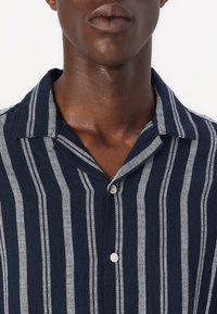 Chemise à rayures bleues marines à manches courtes avec des rayures verticales gris clair, col boutonné et tissu texturé.Elle dispose d'un col standard et de deux boutons.