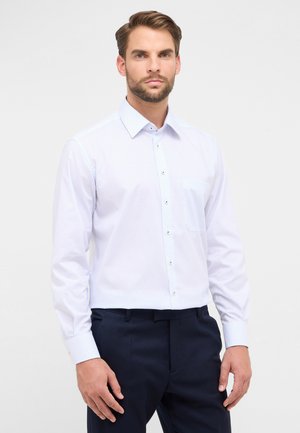 Mann in einem hellblauen karierten Langarmhemd, das in eine dunkelblaue Hose gesteckt ist, steht vor einem schlichten Hintergrund.