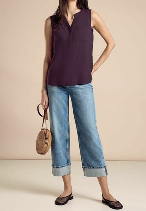 Vrouw die een mouwloze donkerpaarse blouse draagt, brede omgeslagen cropped jeans, zwarte mesh ballerina's, houdt een ronde gevlochten handtas met leren band vast.