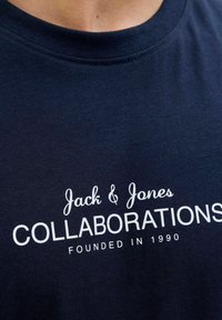 Kara jūras zila kokvilnas T-krekla ar apaļu kakla izgriezumu. Balti burti tekstā rakstīts "Jack & Jones" un "KOLABORĀCIJAS", zemāk norādīts "IZVEIDOTS 1990. GADĀ".