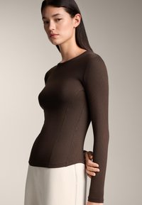 Brown long-sleeve fitted top in ribbed texture, featuring a round neckline and side seam detailing, paired with light cream trousers.