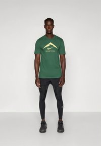 Grønn Nike Trail T-skjorte med gul logo, kombinert med svarte leggings og treningssko, designet for utendørsaktiviteter.