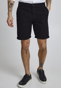 Schwarze Baumwollshorts mit umgeschlagenen Säumen, seitlichen Taschen und Knopfverschluss. Getragen mit schwarzen Sneakers vor einem neutralen Hintergrund.