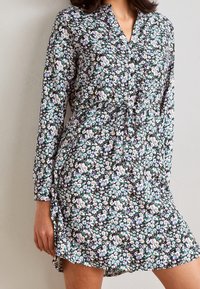 Vestido negro de flores con mangas largas, un cuerpo abotonado, cintura con cordón y una forma fluida. Presenta patrones de flores en morado, blanco y verde.