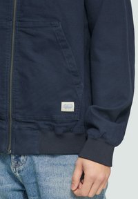 Veste zippée marine avec des poignets et un ourlet côtelés, présentant des poches latérales et une petite étiquette de marque rectangulaire sur le devant gauche.
