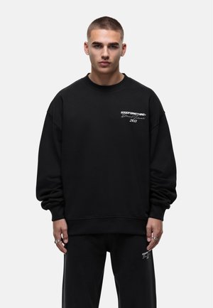 Sweatshirt noir avec un col rond, des épaules tombantes et un texte brodé blanc sur la poitrine, associé à un pantalon de survêtement noir.