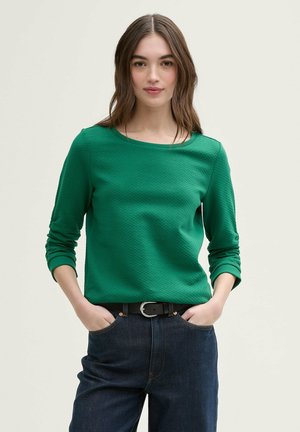 Grön strukturerad långärmad topp med rundad halsringning, kombinerad med mörka denimjeans och ett svart bälte med en silverfärgad spänne.