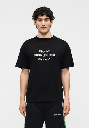Mladý muž s kučeravými vlasmi nosiace čierne tričko s bielym textom "Budú ťa ignorovať, kým nebudú môcť" a čierne nohavice.